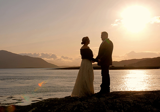 Bride and groom hold hands at shoreline with sunset in background